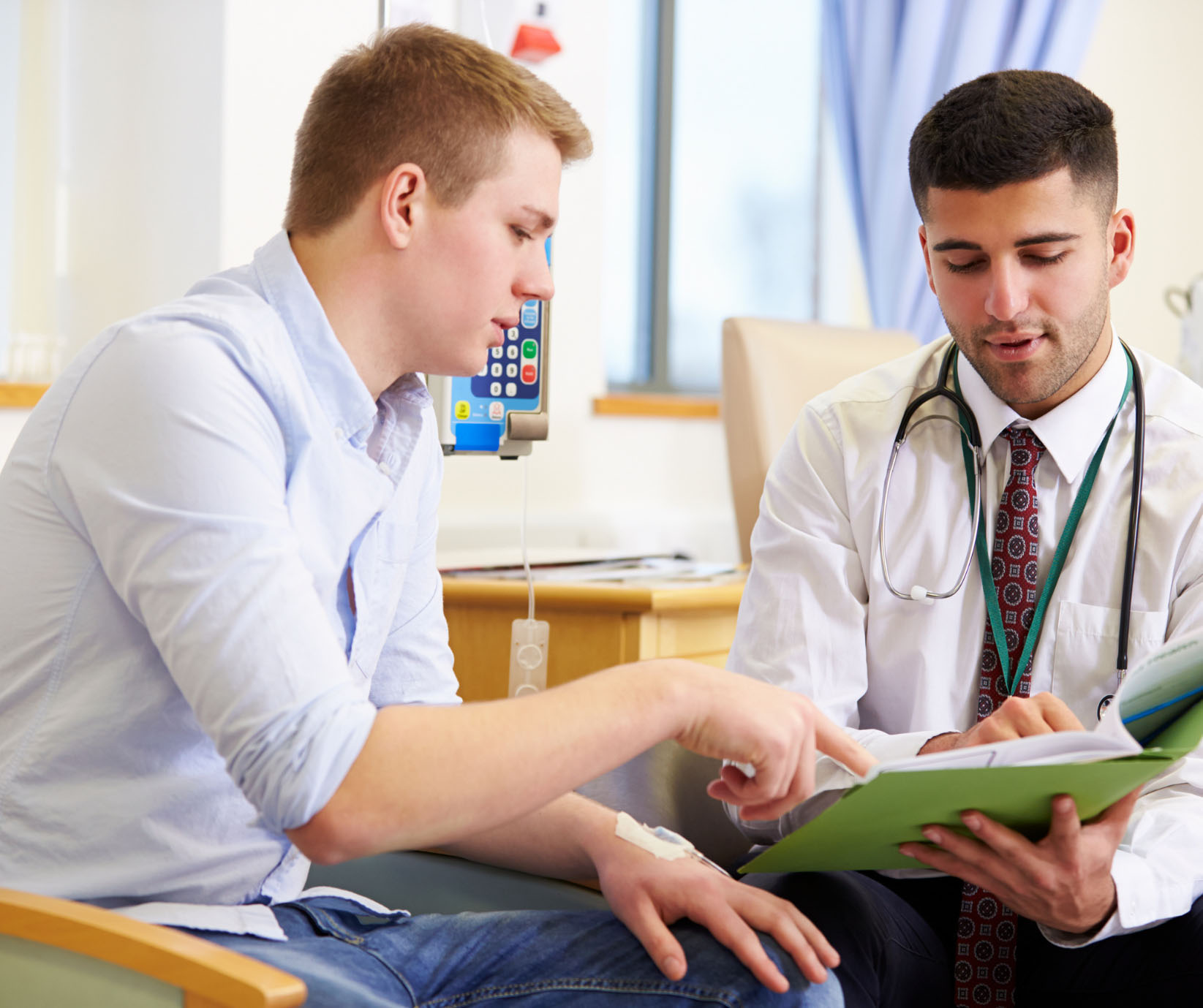 A young male patient with an IV drip in his arm sits talking with a doctor, pointing to documents the doctor is holding in a green folder during a consultation. The scene takes place in a medical office or infusion room.
