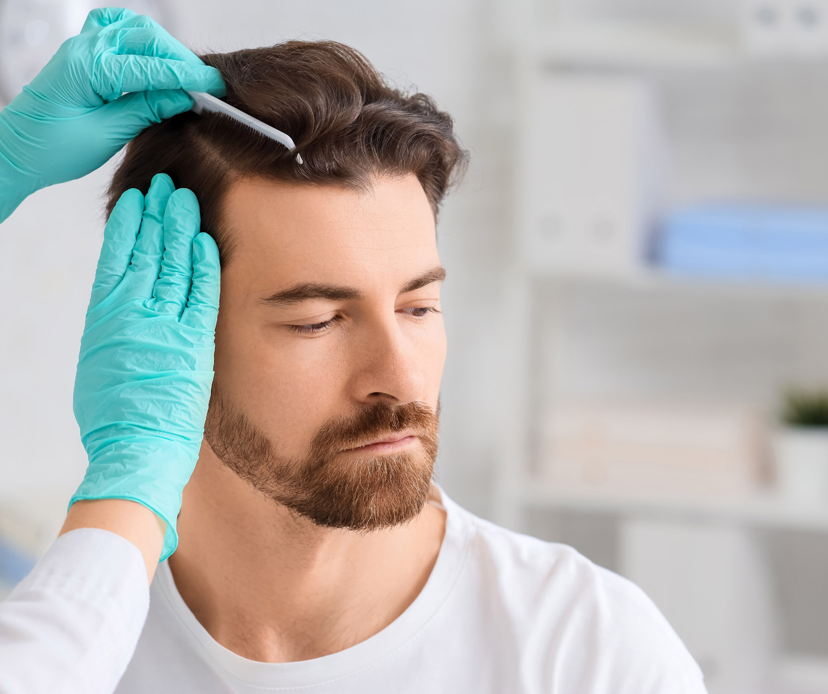 A bearded man receives a trichology or dermatological examination as a technician in blue gloves applies liquid treatment with a fine-toothed comb to his hair.