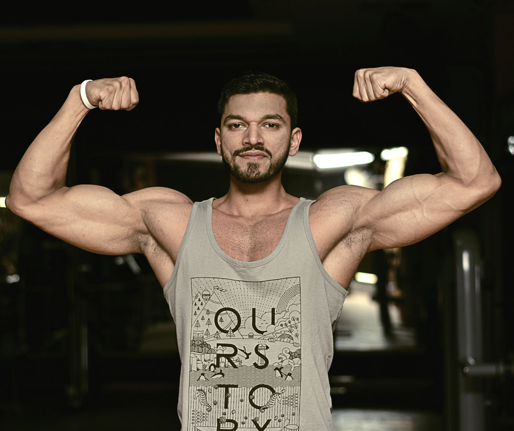 A muscular man in a grey tank top flexes his biceps symmetrically, staring intensely at the camera in a dimly lit gym environment. He has a slight beard and wears a white wristband on his left arm.
