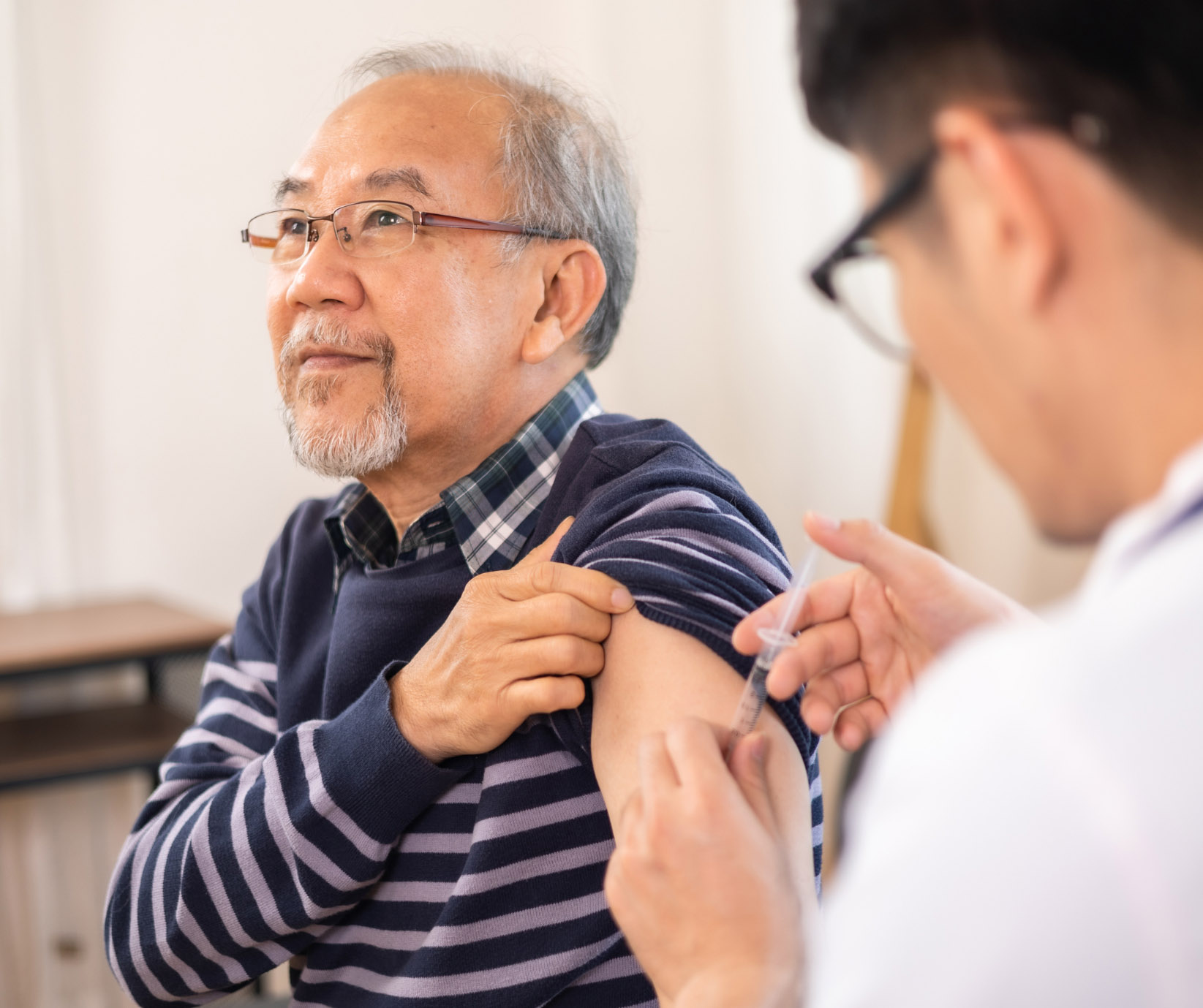 An elderly Asian man receives an injection or vaccine in his upper arm from a doctor, pulling up the sleeve of his striped sweater. The man looks attentive while the healthcare worker, seen from the back, administers the shot.