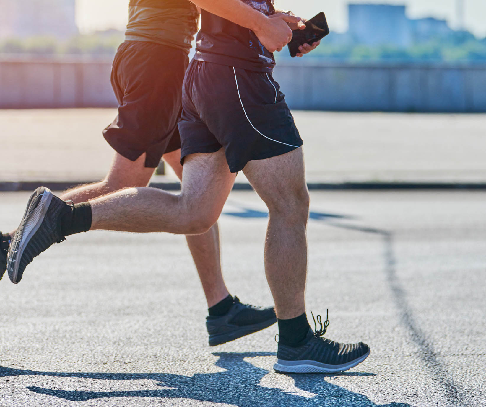 Close-up of the lower bodies of two runners in black shorts and sneakers jogging outdoors on a sunny day. One runner is holding a black smartphone or tablet while exercising.