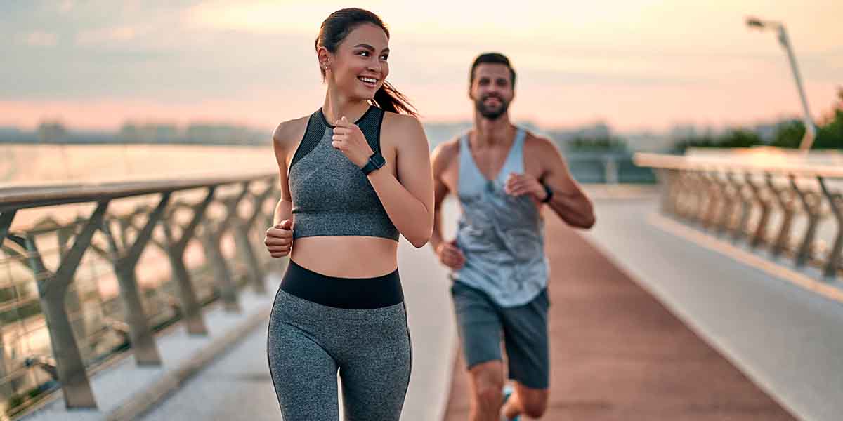 A woman and man joyfully jog on a waterfront path at sunrise, both wearing fitness attire - https://www.coremenshealth.com/